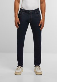 Tumši zila džinsu auduma bikses ar šauru piegriezumu, kurām ir standarta piecu kabatu dizains un smalkas šuvju detaļas. Valkātas ar cieši pieguļošu baltu topi.