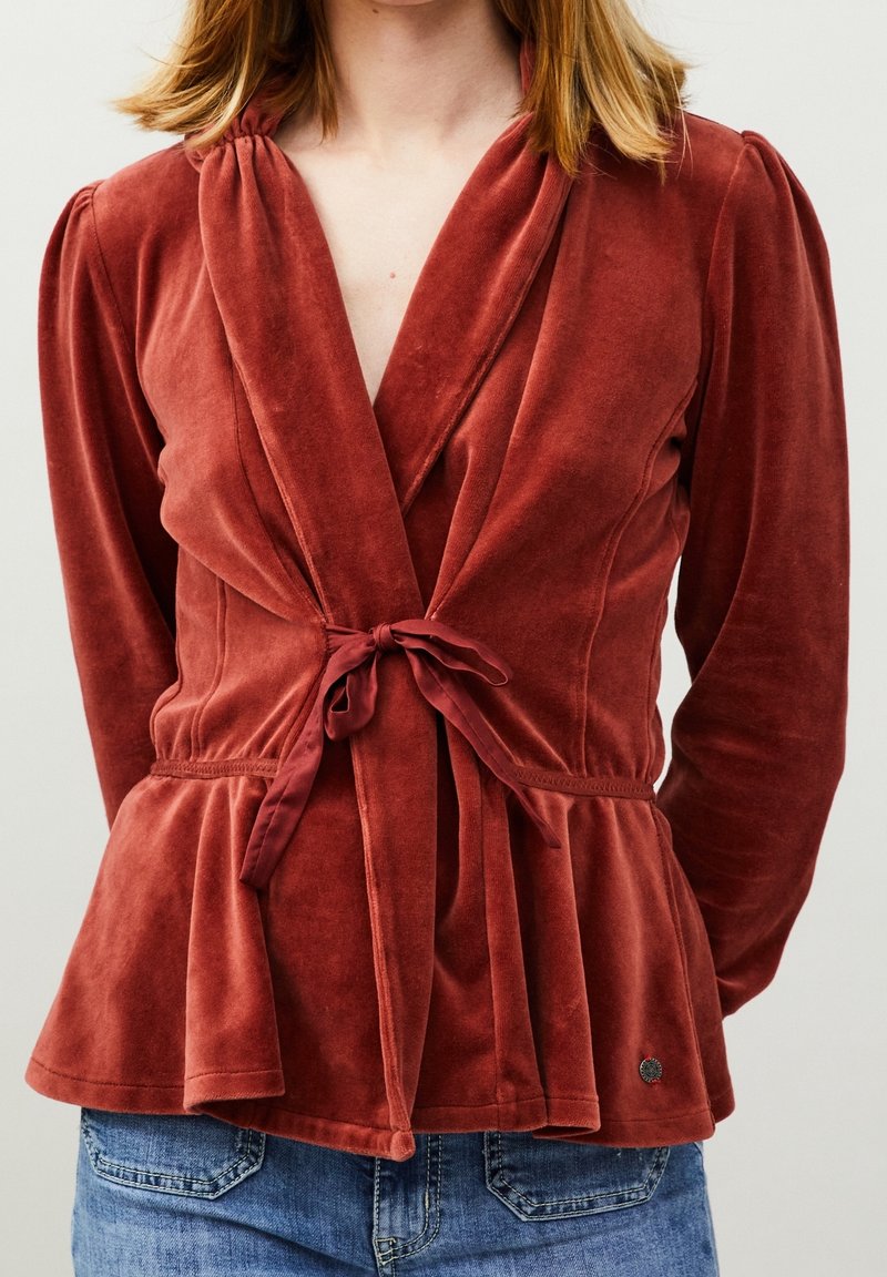 Top de terciopelo suave color óxido, con mangas largas, detalle de pliegues y un lazo en la cintura. Presenta una forma de peplum y un pequeño logo en el dobladillo.