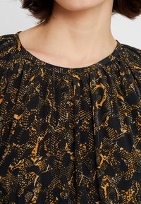 Femme portant un chemisier noir à coupe ample avec un imprimé animalier abstrait aux tons marron et beige.