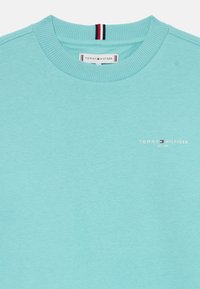 Lyseblå crewneck sweatshirt med ribbestrikket krave, lille Tommy Hilfiger-logo og tekst på brystet, samt rød, hvid og blå stribedetalje indeni kraven.