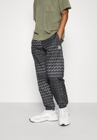Svarta Adidas joggingsbyxor har ett upprepande logomönster, elastiska muddar och sidofickor. En texturerad olivgrön t-shirt bärs ovanpå.