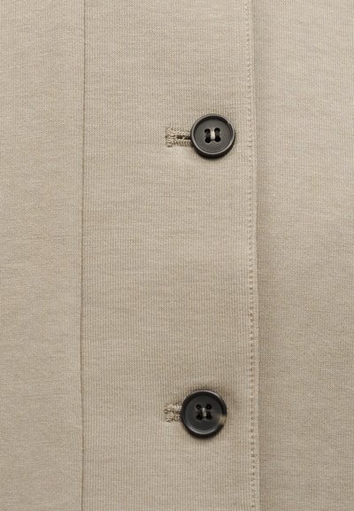 Μπεζ πλεκτό ύφασμα με κάθετη ραφή και δύο μαύρα κουμπιά με τέσσερις τρύπες τοποθετημένα σε κάθετη γραμμή.