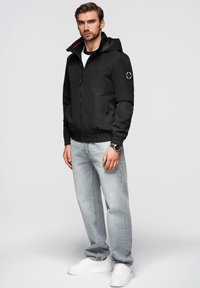 Sort hættetrøje med lynlås foran, elastiske manchetter og et logo-patch på ærmet. Båret med lysegrå jeans og hvide sneakers.