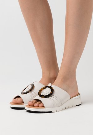 White slide sandals with a textured leather upper, featuring two large tortoiseshell circular accents on the front and a lightweight sole.