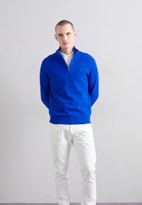 Modra jopica z zipom in rebrastim materialom, z teksturiranim dizajnom. V kombinaciji z belimi jeans hlačami. Model stoji pred enobarvno ozadje.