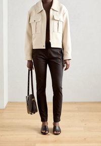 Krémová zkrácená bunda s dvojitými náprsními kapsami, černé úzké kalhoty, černá taška a černé pantofle. Hladké textury a přizpůsobený střih.
