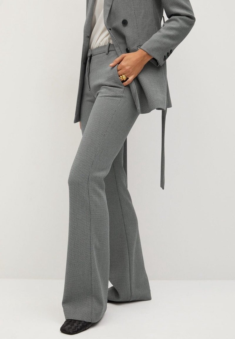 Femme portant un pantalon large gris texturé avec un blazer assorti sur un haut blanc, des chaussures noires tissées et des bagues en or à la main.