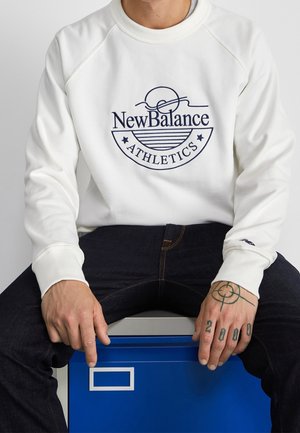 Osoba ubrana w białą bluzę New Balance Athletics i ciemne dżinsy, siedząca na niebieskiej szufladzie, ręce spoczywające na kolanach z widocznymi tatuażami.