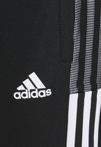 Černé sportovní kalhoty s bílým logem Adidas. Texturovaná tkanina má na boku tři bílé vertikální pruhy.