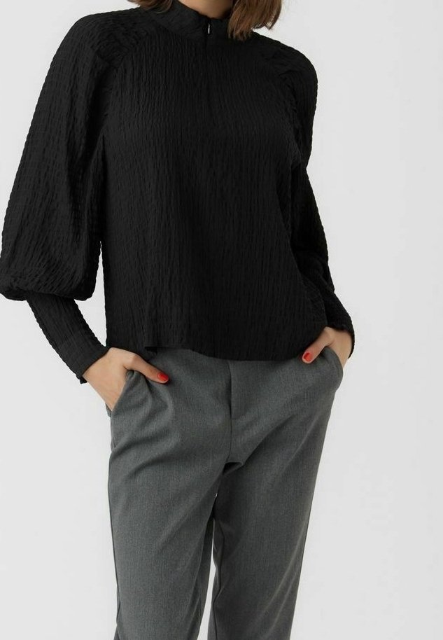 Femme portant un chemisier noir texturé à manches longues avec des épaules bouffantes et un pantalon gris sur mesure, les mains dans les poches.