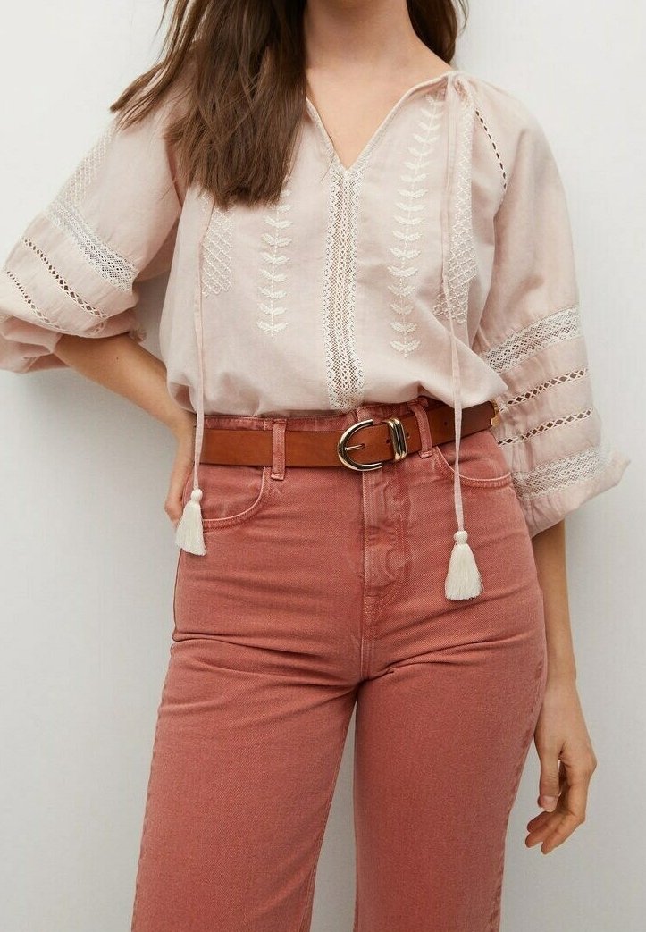 Blouse rose clair avec broderie florale et manches ouvertes, associée à un jean taille haute rose clair et une ceinture marron avec une boucle ronde.