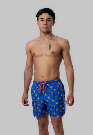 Giovane uomo in piedi, senza maglietta, indossa pantaloncini da bagno blu con stampa colorata di uccelli tropicali e coulisse arancione, sfondo grigio neutro.