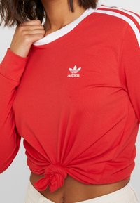 Röd långärmad tröja med vit ribbad krage och vita sidostripes, med en knuten detalj vid midjan och en broderad logotyp.