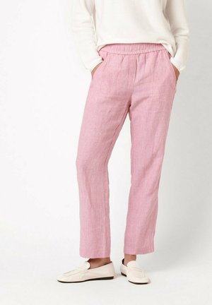 Person wearing light pink linen pants with elastic waist and white loafers, hands in pockets, against plain background.