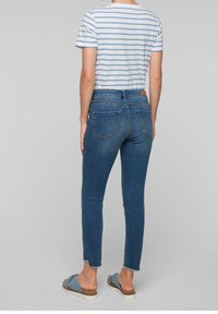 Blårandig kortärmad skjorta, åtsittande blå denimjeans med trasig fåll och ljusblå sandaler med brun sula, sedd bakifrån.