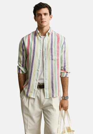 Jonge man met een multicolor verticaal gestreept shirt, witte broek, polshorloge, en een beige draagtas vasthoudend tegen een effen achtergrond.