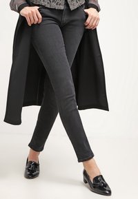 Svarta skinny jeans kombinerat med en mörk, flödande kappa. Modellen bär svarta glänsande loafers med en dekorativ tofs.