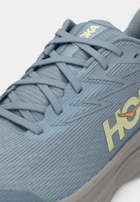 Αθλητικό παπούτσι ανοιχτού γαλάζιου με υφασμάτινη υφή, δεσίματα σε σχηματισμό σταυρού και κίτρινο λογότυπο "HOKA" με πορτοκαλί λεπτομέρεια στο πλάι.