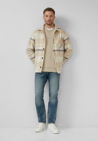 Béžová károvaná bunda s čiernymi akcentmi, zapínanie na gombíky a vreckom na hrudi, v kombinácii so svetlobéžovým mikinou, modrými džínsy a bielymi teniskami.