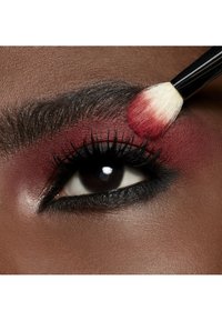 Gros plan sur un œil avec un fard à paupières rouge profond et un eyeliner noir épais. Un pinceau à maquillage ajoute de la couleur avec des poils doux contre une peau lisse.