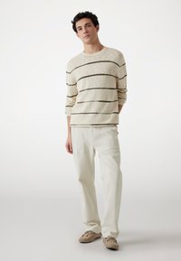 Pull léger beige avec des rayures horizontales marron foncé, associé à un pantalon en velours côtelé écru. Les chaussures sont en daim marron avec des accents en lacets.