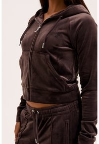 Femme portant un sweat à capuche zippé en velours marron foncé avec poches avant et pantalon assorti à cordon avec poches latérales zippées.