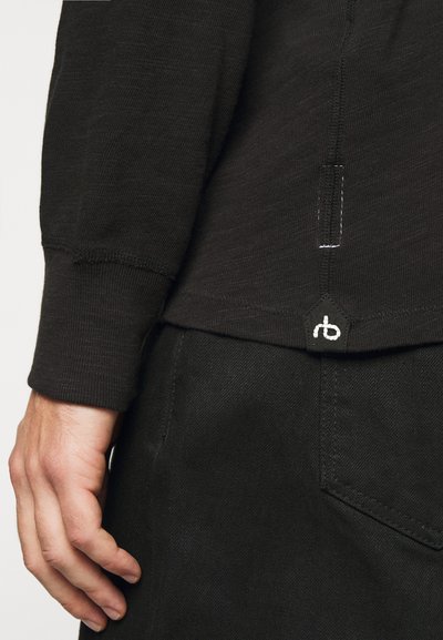 Black long-sleeve shirt featuring a textured fabric, visible stitching, and a small logo patch at the hem with a back detail.