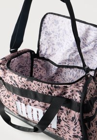 Ανοικτό ροζ και μαύρο σπορ τσάντα ταξιδιού με μαύρα λουριά και μεγάλο λευκό λογότυπο Puma στο πλάι.