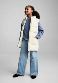 Gilet in piuma bianco sopra un maglione blu a trama, abbinato a jeans azzurri a gamba larga e scarpe slip-on nere. Sfondo neutro.