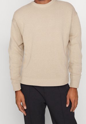 Pull en tricot beige avec un col rond et des manches longues. L'ourlet et les poignets présentent des détails côtelés, créant une finition texturée.