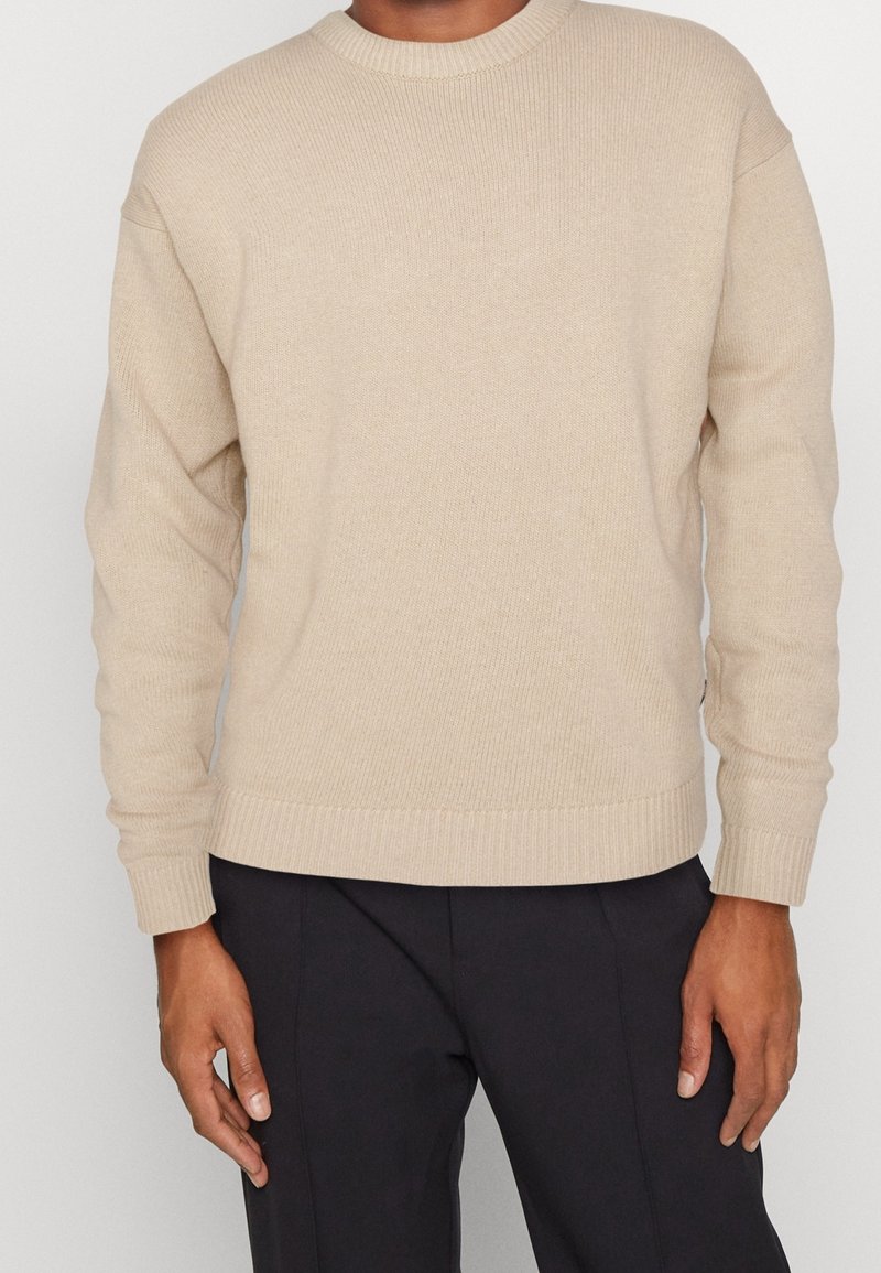 Pull en tricot beige avec un col rond et des manches longues. L'ourlet et les poignets présentent des détails côtelés, créant une finition texturée.