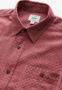 Chemise à motifs rouges avec un col boutonné, dotée d'une poche poitrine et de boutons marron. Le matériau a une finition texturée.