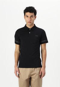 Čierna polo košeľa vyrobená z mäkkej tkaniny, s golierom, krátkymi rukávmi a logom "GUESS" na hrudi a na rukáve v kontrastnej sivej farbe.