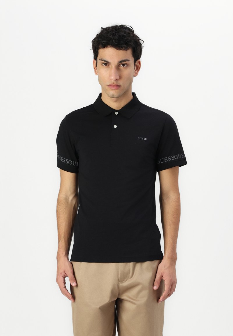 Čierna polo košeľa vyrobená z mäkkej tkaniny, s golierom, krátkymi rukávmi a logom "GUESS" na hrudi a na rukáve v kontrastnej sivej farbe.