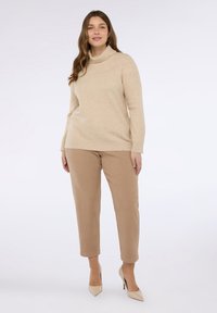 Kobieta w beżowym swetrze z golfem, jasnobrązowych spodniach  cropped i beżowych szpilkach stoi na tle białym.