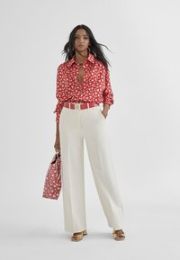 Blusa a motivi rossi e bianchi con scollatura a V profonda, pantaloni a gamba larga color crema, una cintura rossa abbinata e una borsa a tracolla con motivo. Sandali dorati.