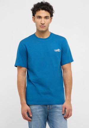 Mężczyzna z kręconymi włosami, ubrany w niebieską koszulkę Mustang i jasnoniebieskie jeansy, stojący na jednolitym jasnym tle.