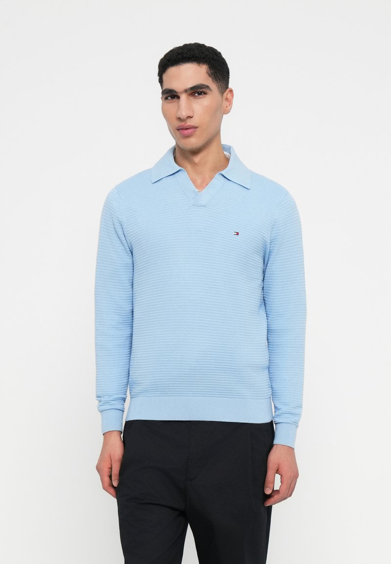 Férfi világoskék bordázott hosszú ujjú pulóverben, gallérral és fekete nadrágban, egy sima fehér háttér előtt áll.