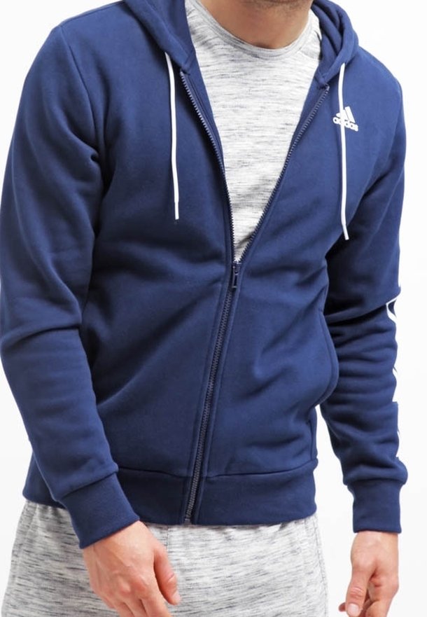 Homme portant un sweat à capuche zippé Adidas bleu marine par-dessus un t-shirt gris clair et un pantalon gris assorti, debout les mains près des hanches.