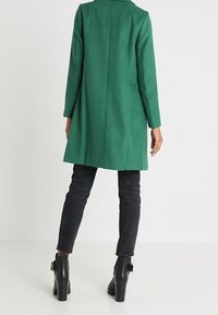 Abrigo verde de mezcla de lana con silueta recta, cuello con muesca y mangas largas, combinado con jeans ajustados negros y botines de tacón.