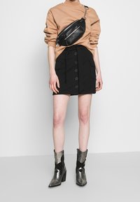 Jupe noire boutonnée sur le devant avec poches latérales, associée à un sweat-shirt marron clair et des bottines métalliques à surfaces texturées.