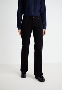 Black cotton trousers with a straight leg design, featuring front buttons and pockets. Worn with a dark denim shirt and black shoes.