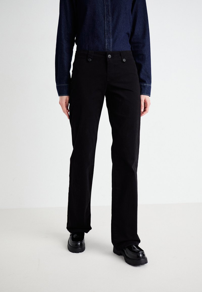 Black cotton trousers with a straight leg design, featuring front buttons and pockets. Worn with a dark denim shirt and black shoes.