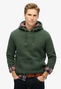 Felpa verde con tasca frontale e cordini, sovrapposta a una camicia a quadri. Tessuto testurizzato, cuciture visibili e un accento con logo.