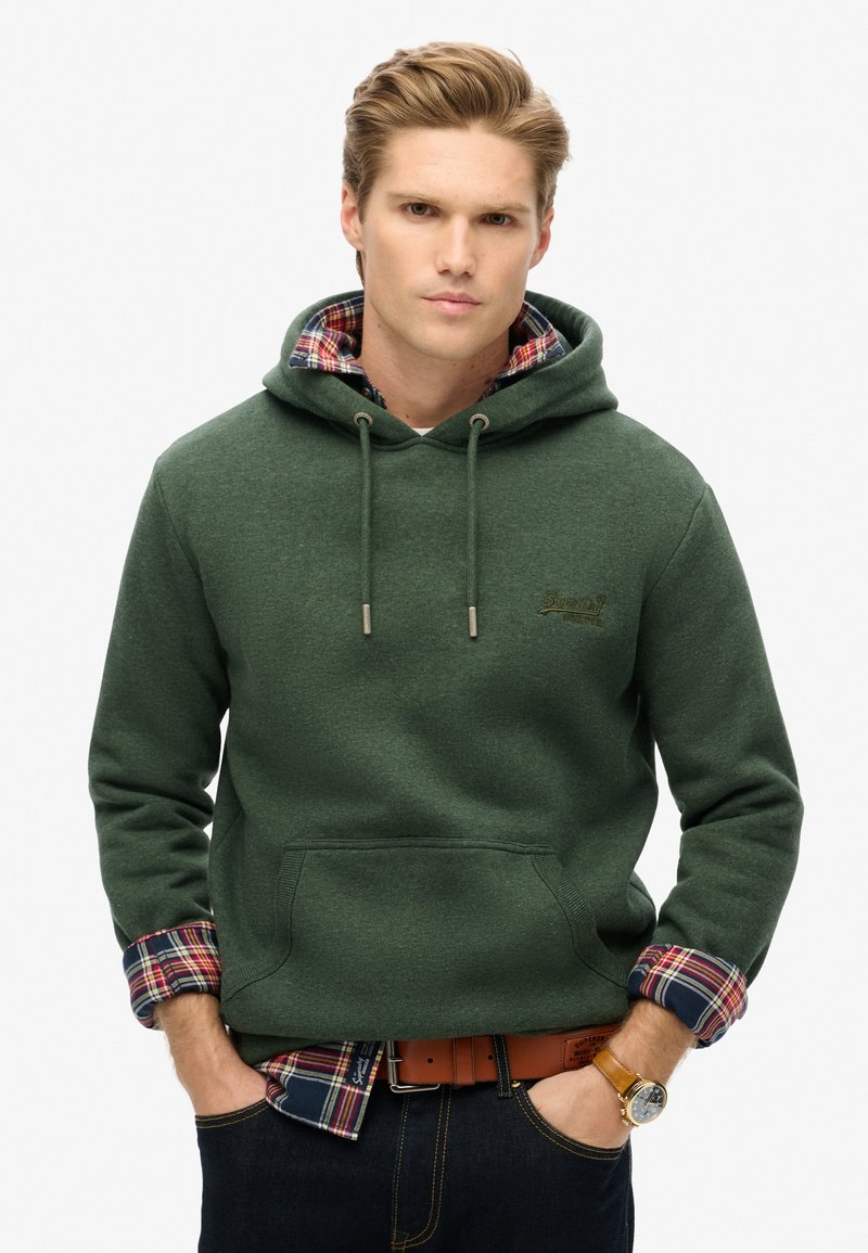 Felpa verde con tasca frontale e cordini, sovrapposta a una camicia a quadri. Tessuto testurizzato, cuciture visibili e un accento con logo.