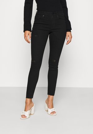 Vrouw die zwarte gescheurde skinny jeans draagt en witte sandalen met hoge hakken en geknoopte bandjes, staand tegen een lichte achtergrond.