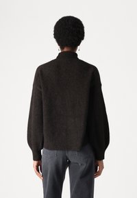 Tummanruskea, ylisuurikokoinen neulepusero, jossa korkea kaulus, resoriset hihansuut ja helma. Pehmeä, teksturoitu kangas ja hieman ilmavat hihat.