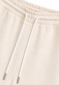 Gros plan de la taille d’un pantalon de jogging beige avec des cordons blancs dotés d’embouts métalliques.