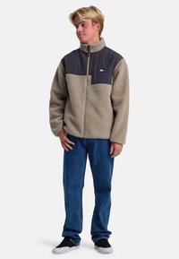 Chaqueta de forro polar con un cuerpo beige, panel superior gris y cremallera frontal. Presenta un parche con logo, bolsillos laterales y un collar alto. Combinada con vaqueros azules.