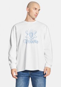 Biała koszulka z długim rękawem z jasnoniebieską grafiką przedstawiającą herb oraz ozdobny tekst z przodu. Luźny krój, miękki bawełniany materiał.